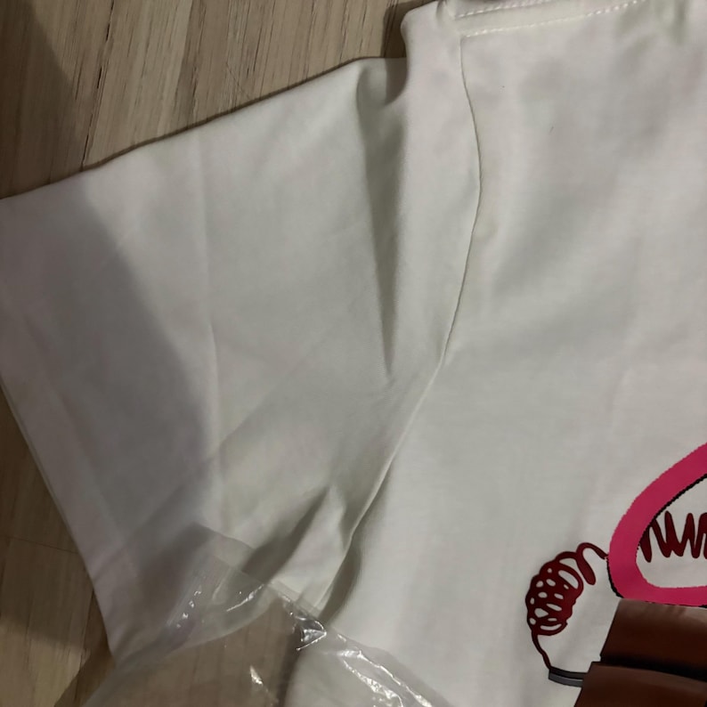 May include: White t-shirt with a graphic design. The design features a cartoon image with pink and brown accents. The shirt is folded and partially covered in a clear plastic bag.