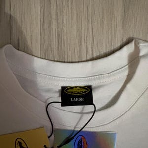 May include: Close-up of a white t-shirt collar with a black tag that reads "LARGE". The tag has a gold emblem. A second tag with a holographic design is visible.