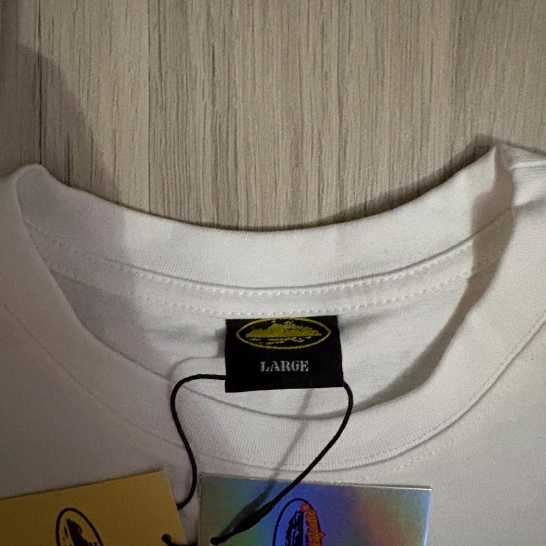 May include: Close-up of a white t-shirt collar with a black tag that reads "LARGE". The tag has a gold emblem. A second tag with a holographic design is visible.