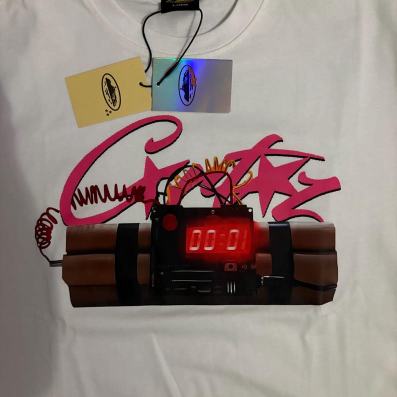 May include: White t-shirt featuring a pink graphic of the word "Crazy" with a bomb design. The bomb has a digital display showing "00:01". The shirt has two tags attached.