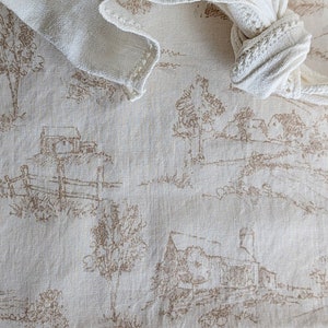 May include: A white linen kitchen towel with a delicate crocheted edge is folded and tied into a knot. The towel is lying on a cream-colored fabric with a repeating pattern of brown farmhouses and trees.