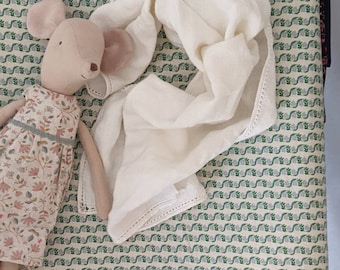 Op de afbeelding: Een zachte, beige muispop met een bloemenjurk, naast een crèmekleurige linnen sjaal. De items liggen op een stof met patronen met groene en grijze bloemmotieven.