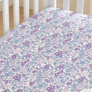May include: A white wooden crib features a fitted sheet with a dense floral pattern in shades of purple, light blue, and soft pink. A cream-colored knit blanket is draped over the front rail of the crib, which has vertical slats.