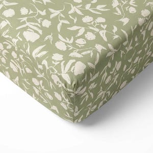 May include: A fitted sheet with a sage green background and a repeating pattern of off-white floral and leaf designs. The fabric is folded over, showing the corner seam. The sheet is likely for a bed.