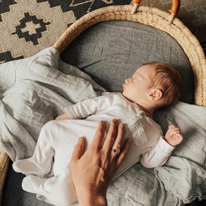 May include: A baby sleeping in a woven basket with a gray and white striped blanket. A hand is gently resting on the baby's chest.