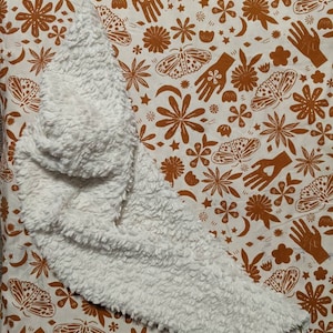 May include: A white, fluffy, sherpa-like blanket is draped over a cream-colored fabric. The fabric has a repeating pattern of brown floral and celestial designs, including hands, moths, flowers, stars, and crescent moons.