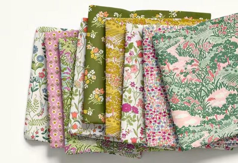 May include: A collection of folded fabric pieces featuring various floral and landscape patterns. The fabrics display a range of colors, including pink, green, yellow, and blue, with detailed botanical designs. The fabrics are likely for sewing or crafting.