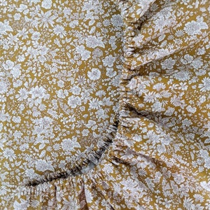 May include: Close-up of a fitted sheet with a floral pattern. The sheet features a mustard yellow background covered in small white flowers. The edges are gathered, creating a ruffled effect.