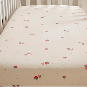 May include: A cream-colored fitted crib sheet with a repeating pattern of pink strawberries and small pink dots. The strawberries have green leaves and are scattered across the sheet.