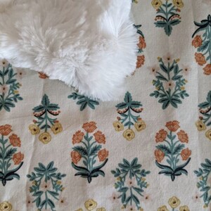 May include: A white, fluffy, faux fur item rests on a cream-colored fabric with a repeating floral pattern. The pattern features stylized flowers in shades of orange, yellow, and pink, with teal green leaves and stems.