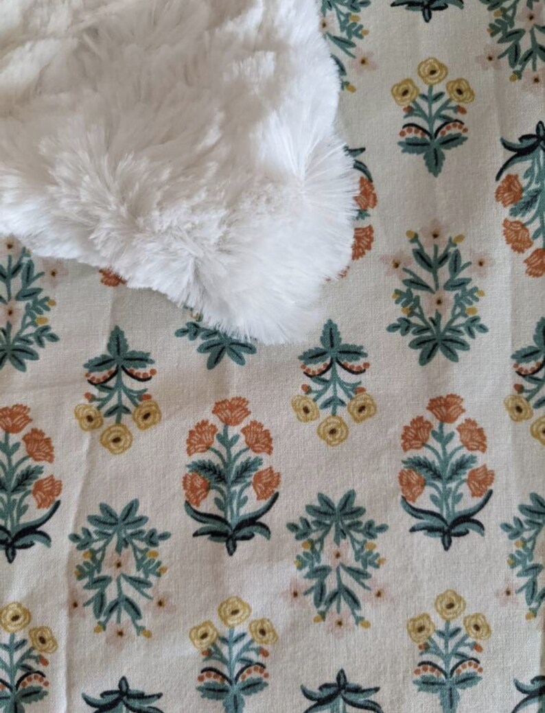 May include: A white, fluffy, faux fur item rests on a cream-colored fabric with a repeating floral pattern. The pattern features stylized flowers in shades of orange, yellow, and pink, with teal green leaves and stems.