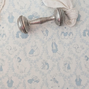 Puede incluir: Un sonajero de bebé de plata con una superficie texturizada. El sonajero está sobre una tela blanca con un patrón azul y blanco de conejos, patos y otros animales.