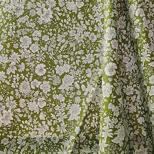 May include: Close-up of a fabric with a floral pattern. The fabric is a deep olive green color, covered in a repeating pattern of white flowers and leaves. The fabric is draped, showing folds and texture.