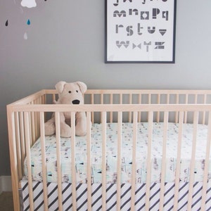 May include: A wooden crib with a white and black striped fitted sheet. A stuffed dog toy is sitting in the crib. The crib is in front of a gray wall with a black framed print of the alphabet hanging above it.