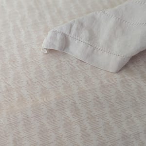 May include: A close-up of a cream-colored bed sheet with a subtle, repeating pattern of diagonal lines in a light brown hue. A folded corner of a matching pillowcase is visible, with delicate stitching along the edge.