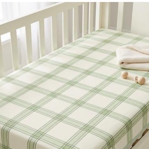 May include: A fitted crib sheet featuring a light green and cream plaid pattern. The sheet is stretched over a mattress and includes a folded cream-colored knit blanket and a small, light-colored wooden rattle resting on the surface.