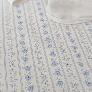 May include: Close-up of a fabric with a repeating floral pattern. The fabric features vertical stripes in light blue and white, with delicate blue floral sprigs and dotted accents. A white fabric edge is visible in the upper portion of the image.