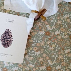 May include: A white linen napkin held by a wooden napkin ring, resting on a floral patterned fabric. A book with the title "A Trip to the Forest" and a pinecone illustration is visible.