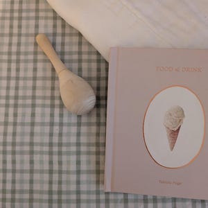 May include: A light pink hardcover book titled FOOD & DRINK by Tabitha Paige with an ice cream cone illustration, a smooth light-colored wooden rattle, and a white fabric piece resting on a sage green and white checkered fabric surface.