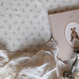 May include: A white crocheted blanket with a lace trim, a pink book with the word "TOYS" on the cover, and a silver rattle. The book has a picture of a teddy bear on the cover.