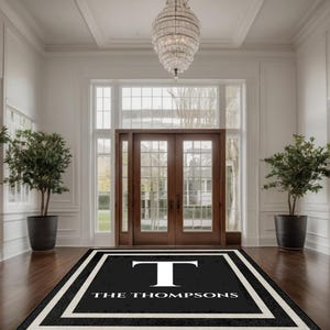Può includere: Un zerbino personalizzato in bianco e nero con il nome "THE THOMPSONS" e una grande lettera "T" bianca. Il tappeto è rettangolare e si trova su un pavimento in legno scuro in un ingresso ben illuminato con un grande lampadario.