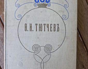 May include: A vintage book with a light beige cover. The cover features a blue floral design above a circular frame with the Cyrillic text "В.Н. ТЮТЧЕВЪ". The book's title is in a dark font. The book is on a wooden surface.