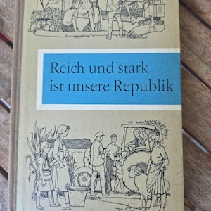 Puede incluir: Libro de tapa dura vintage con una cubierta beige y un cuadro de texto azul. La cubierta presenta ilustraciones en blanco y negro de niños, equipos de construcción y escenas de granja. El título "Reich und stark ist unsere Republik" está impreso en la cubierta.