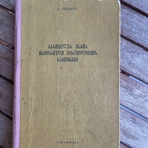May include: A hardcover book with a muted olive-green cover and a light pink fabric spine. The front cover features black text in the Georgian script, including the title and author name, with the word "განათლება" printed at the bottom.