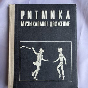 May include: A black hardcover book with a light beige spine. The cover features white text reading "РИТМИКА МУЗЫКАЛЬНОЕ ДВИЖЕНИЕ" above a white-lined rectangular frame containing a graphic illustration of a girl holding a ribbon and a boy dancing.