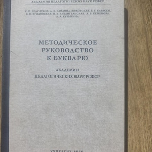 Op de afbeelding: Een vintage boek in het Russisch, getiteld "METOДИЧЕСКОЕ РУКОВОДСТВО К БУКВАРЮ". De omslag is lichtgrijs met zwarte tekst, inclusief de naam van de uitgever en het jaar 1956. Het boek is van de Academie voor Pedagogische Wetenschappen van de RSFSR.