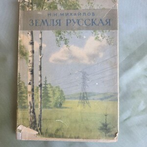 以下が含まれることがあります： N.N.ミハイロフ著「ЗЕМЛЯ РУССКАЯ」というタイトルのヴィンテージのハードカバー本。表紙には、白樺の木、緑の野原、青い空の下にある送電塔の風景イラストが描かれています。本の表紙の端には摩耗が見られます。