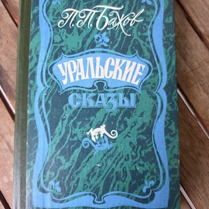 May include: A hardcover book with a dark green and blue marbled cover featuring a decorative border. The text reads P.P. Bazhov and Uralskie Skazy in Cyrillic script. A small white and blue lizard illustration is centered near the bottom of the cover.