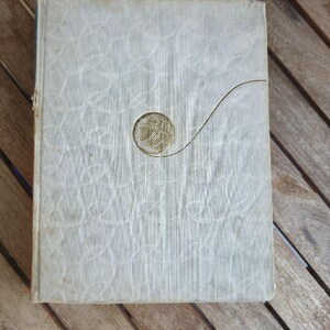 May include: A rectangular hardcover book with a textured off-white fabric cover featuring a subtle, embossed swirling pattern. Centered on the front cover is a gold-colored circular graphic depicting a ball of yarn with a single trailing thread.