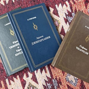 Könnte beinhalten: Drei Hardcover-Bücher mit goldfarbenen Buchstaben und Akzenten sind auf einem gemusterten Textil angeordnet. Die Bücher sind in Grün-, Blau- und Brauntönen gehalten. Jedes Buch hat Text in einer Fremdsprache und ein goldenes Sternmuster.