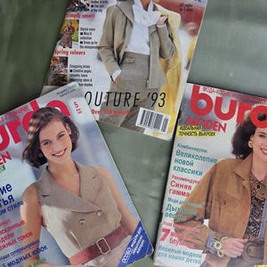 Puede incluir: Se muestran tres revistas de moda vintage de la década de 1990. Las revistas presentan modelos con ropa elegante. Las portadas tienen texto en varios idiomas, incluyendo "OUTURE '93" y "BURDA MODEN". Las revistas están sobre una superficie verde.