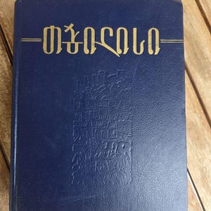 May include: A dark blue hardcover book with a textured leather-like finish. The front cover features gold embossed text in the Georgian script reading "თბილისი" and a subtle, debossed vertical illustration of architectural structures.