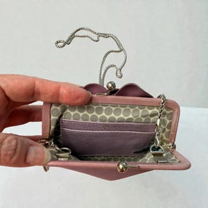Puede incluir: Un bolso de mano de cuero rosa claro con una correa de cadena plateada. El interior presenta un forro con estampado de lunares grises claros y blancos y varias ranuras para tarjetas. El cierre es un cierre de bola plateada.