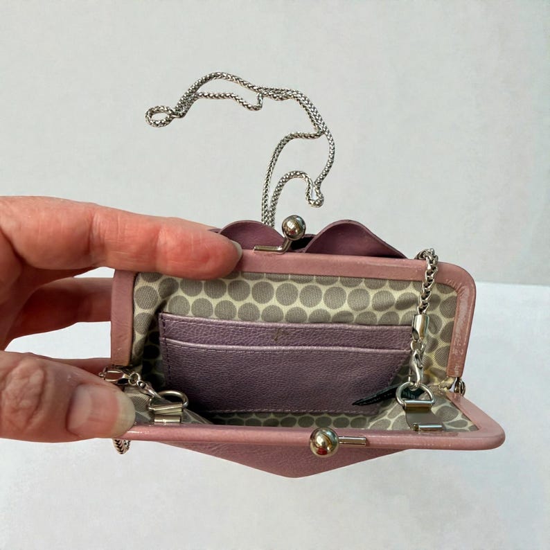 Puede incluir: Un bolso de mano de cuero rosa claro con una correa de cadena plateada. El interior presenta un forro con estampado de lunares grises claros y blancos y varias ranuras para tarjetas. El cierre es un cierre de bola plateada.