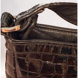 May include: A dark brown handbag with a glossy, embossed crocodile skin texture. The bag features a structured top edge, a smooth interior lining, and a metallic silver-toned hardware ring connecting the handle to the main body of the bag.