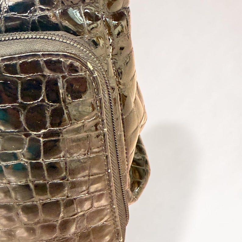 May include: A metallic bronze handbag featuring a glossy, embossed crocodile skin texture. The bag includes a front zippered pocket with a matching metallic zipper pull and a structured, shiny finish that reflects light across its surface.