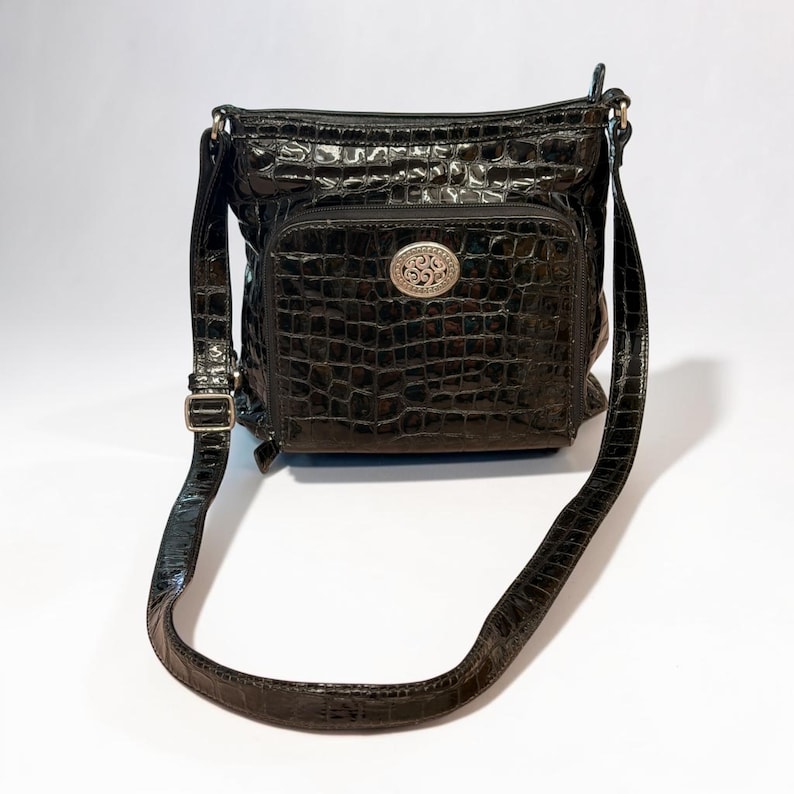May include: A black crossbody bag with a glossy, embossed crocodile skin texture. The bag features a front zippered pocket adorned with a centered, oval-shaped silver-toned metal emblem with an intricate swirling pattern and an adjustable strap.