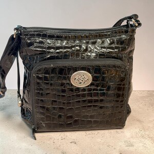 May include: A black crossbody bag with a glossy, embossed crocodile skin texture. The bag features a front zippered pocket adorned with a central oval silver-toned metal emblem featuring intricate swirling patterns and a matching adjustable shoulder strap.
