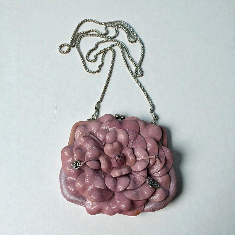 Puede incluir: Un bolso de mano de cuero malva con dise&ntilde;o de flor. El bolso tiene una correa de cadena plateada y un cierre de broche. El dise&ntilde;o floral est&aacute; hecho de p&eacute;talos superpuestos.