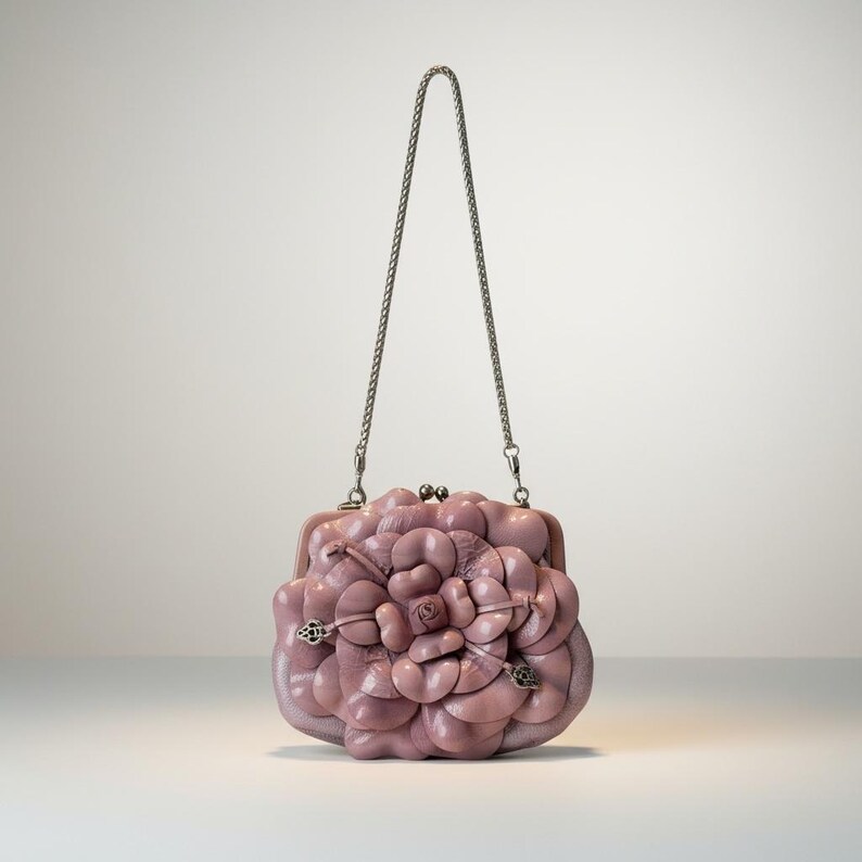 Puede incluir: Un bolso de mano de color malva con un dise&ntilde;o floral. El bolso tiene una correa de cadena plateada y un cierre de broche. El dise&ntilde;o floral est&aacute; hecho de p&eacute;talos en capas, creando un efecto tridimensional. El bolso est&aacute; sobre un fondo blanco.