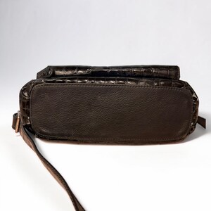 May include: A dark brown handbag featuring a glossy, embossed crocodile skin texture on the upper portion and a smooth, matte finish on the base. The bag includes a thin, adjustable shoulder strap attached to the side with a metallic buckle.