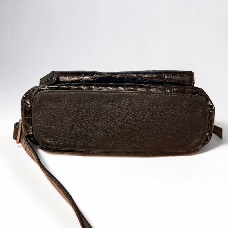 May include: A dark brown handbag featuring a glossy, embossed crocodile skin texture on the upper portion and a smooth, matte finish on the base. The bag includes a thin, adjustable shoulder strap attached to the side with a metallic buckle.