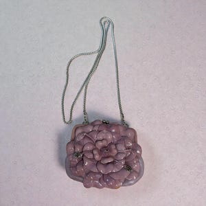 Puede incluir: Un bolso de mano de color lavanda con una cadena plateada. El bolso presenta un gran dise&ntilde;o floral texturizado en la parte delantera y un cierre plateado. Un accesorio &uacute;nico.