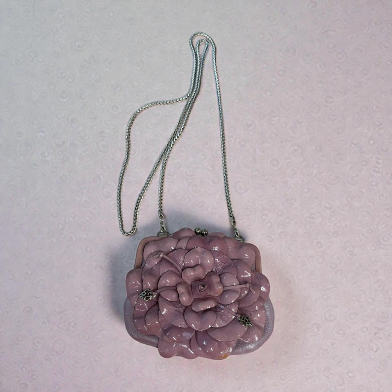 Puede incluir: Un bolso de mano de color lavanda con una cadena plateada. El bolso presenta un gran dise&ntilde;o floral texturizado en la parte delantera y un cierre plateado. Un accesorio &uacute;nico.