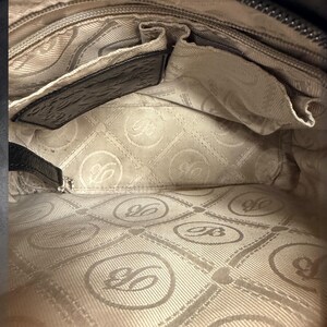 May include: The interior of a handbag featuring a light beige fabric lining with a repeating circular logo pattern containing the letter B. It includes a black leather trim along the top edge, a zippered pocket, and an open slip pocket compartment.