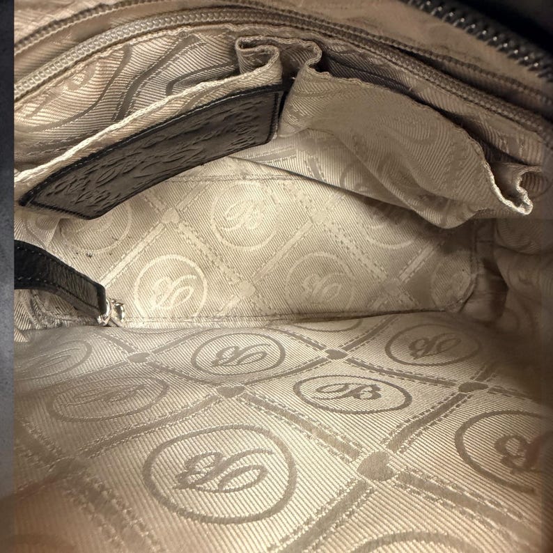 May include: The interior of a handbag featuring a light beige fabric lining with a repeating circular logo pattern containing the letter B. It includes a black leather trim along the top edge, a zippered pocket, and an open slip pocket compartment.